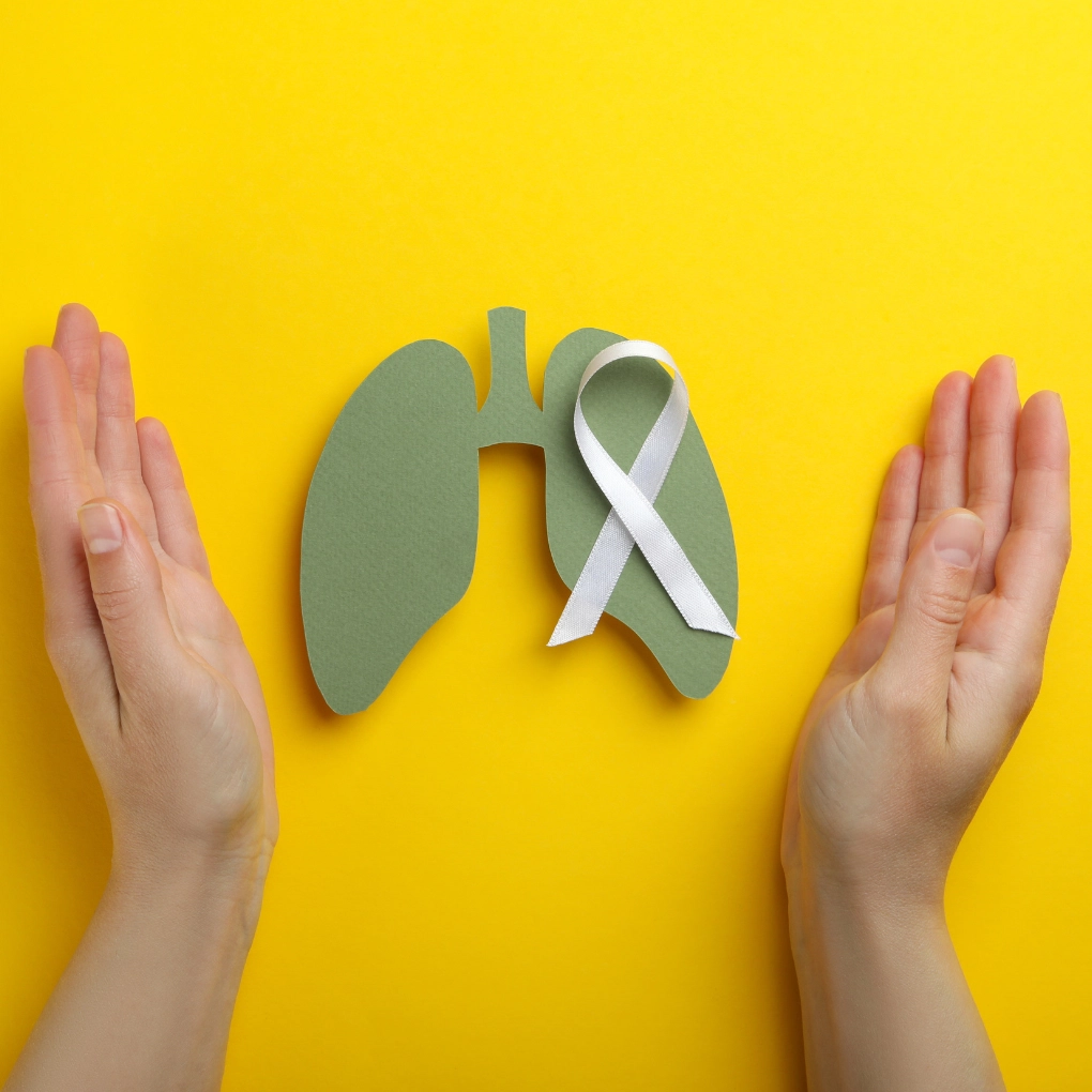 A pair of hands holding a paper lung with a white ribbon on a yellow background. The ribbon is a symbol of hope and support for those who are struggling with cancer.